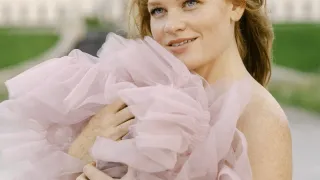 Eine Frau mit blonden Haaren in einem rosa Kleid steht lächelnd vor einem großen Gebäude.