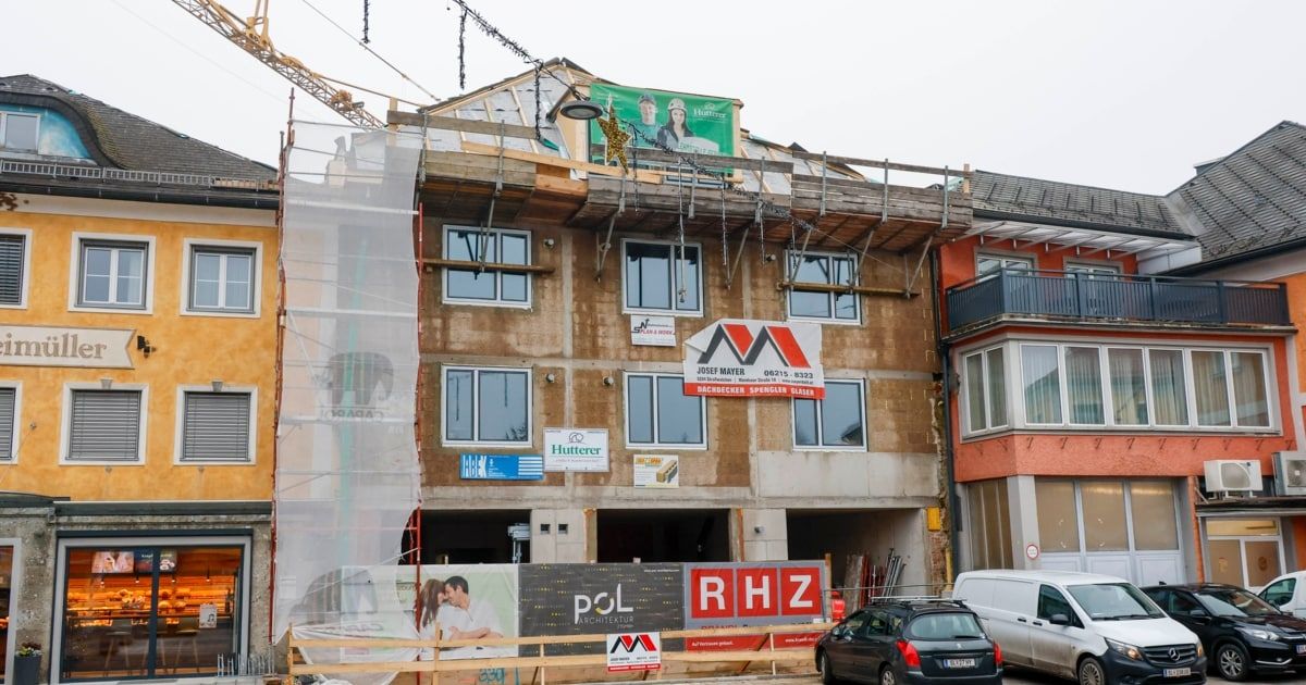 Ein Gebäude im Bau mit mehreren Fenstern, einem Schild und einem Kran oben. Das Gebäude hat ein weißes Banner mit einem Bild eines Paares und einem Text, der 'POL Architektur' lautet. Es gibt geparkte Autos und einen Van an der Seite.