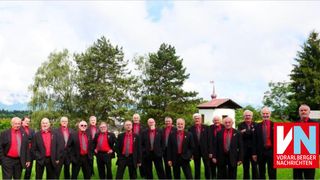 Eine Gruppe von Männern in formeller Kleidung steht auf einer grasbewachsenen Fläche, mit Bäumen und einem kleinen Gebäude im Hintergrund.