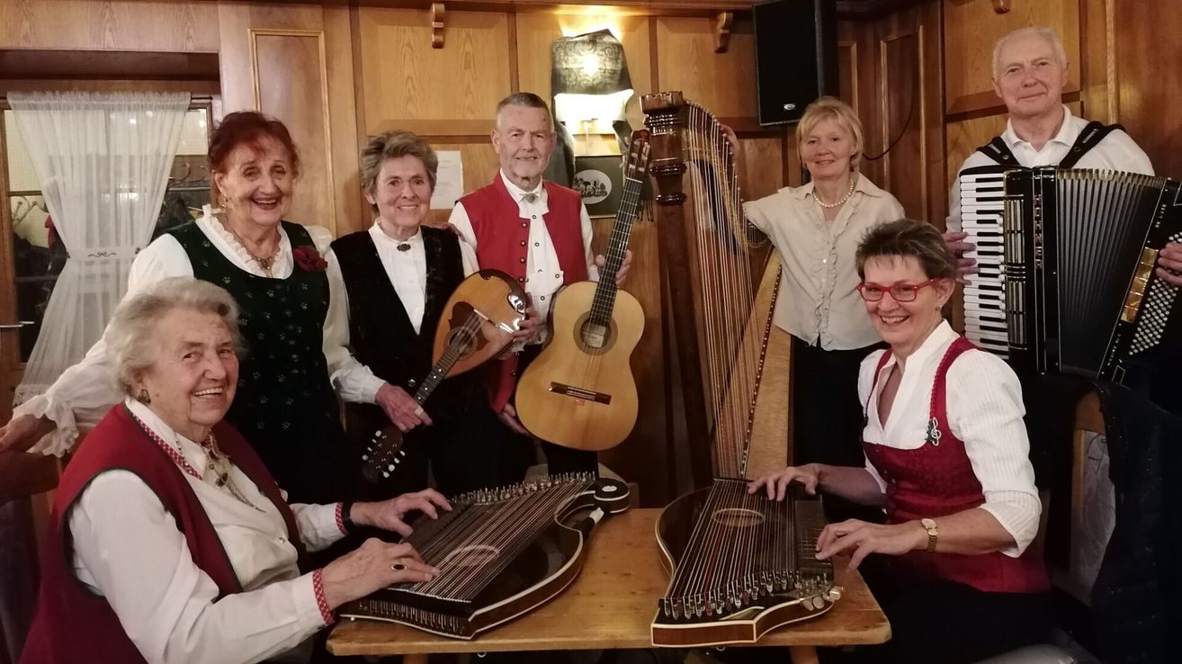Eine Gruppe älterer Erwachsener, gekleidet in traditioneller Kleidung, lächeln und posieren für ein Foto. Sie halten verschiedene Musikinstrumente, darunter Gitarren, Harfen und Zithern, und stehen vor einer Holzwand.