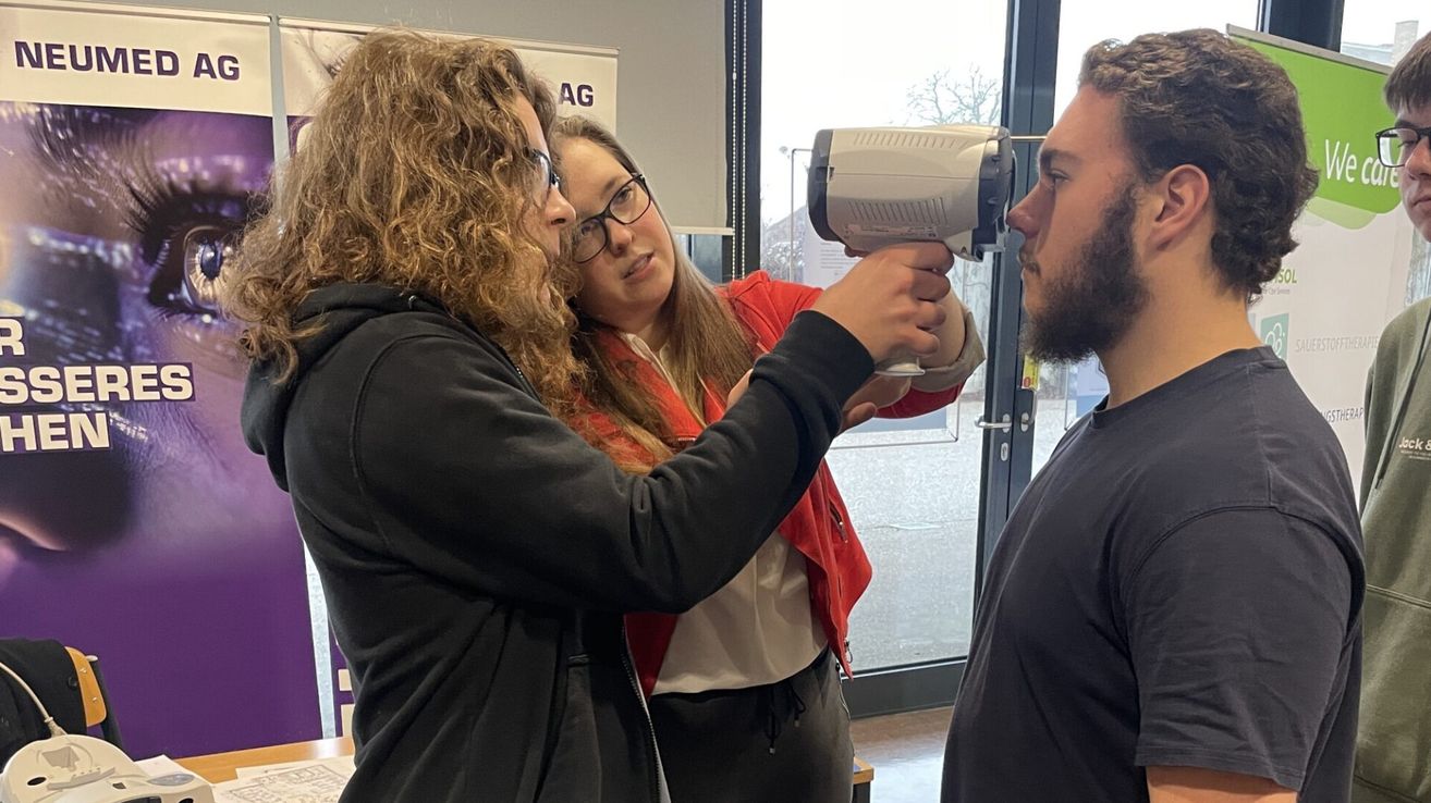 Zwei Frauen mit Brille überprüfen die Temperatur eines Mannes mit einem Thermometer. Der Mann steht mit geschlossenen Augen. Sie befinden sich in einem Raum mit Glastüren und Blick auf Bäume draußen.