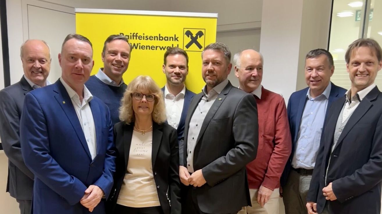 Eine Gruppe von Menschen, möglicherweise ein Team, steht vor einem gelben Banner mit dem Text Raiffeisenbank Wienerwald. Sie alle lächeln und scheinen für ein Foto zu posieren.