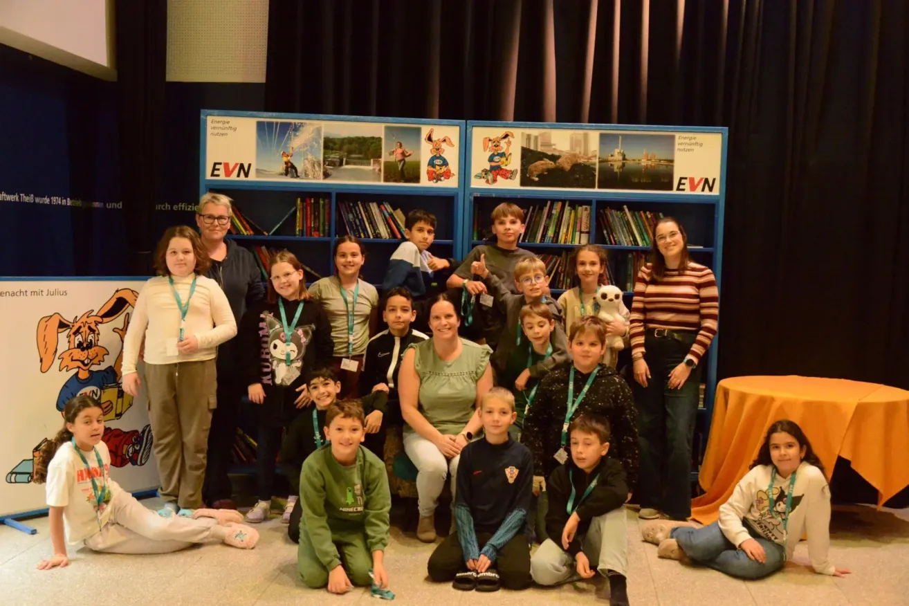 Eine Gruppe von Kindern und Erwachsenen in einem Raum mit blauen Bücherregalen, einer hält einen weißen Teddybär. Zwei Frauen mit Brille und einem Gürtel stehen an den Seiten.