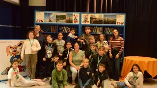 Eine Gruppe von Kindern und Erwachsenen in einem Raum mit blauen Bücherregalen, einer hält einen weißen Teddybär. Zwei Frauen mit Brille und einem Gürtel stehen an den Seiten.
