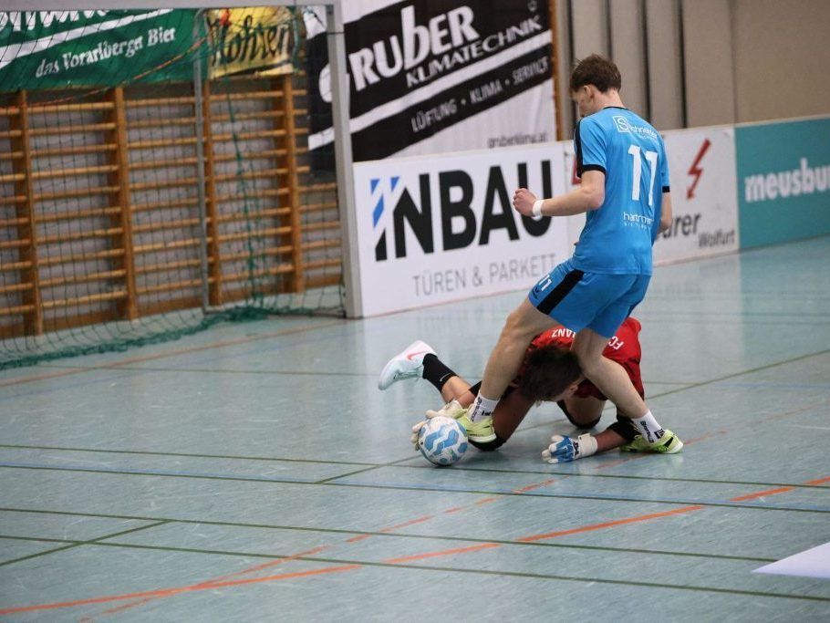 Ein Futsal-Spiel zeigt einen Spieler in Blau, Nummer 17, der auf den Rücken eines roten Spielers tritt und versucht, einen Ball in der Nähe des Tores zu blockieren.