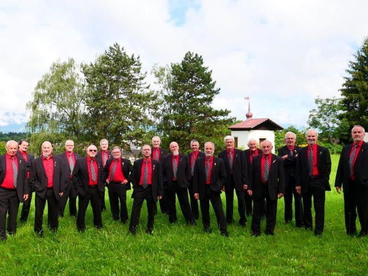 Eine Gruppe von Männern in roten Hemden und schwarzen Anzügen posiert für ein Foto auf einer Wiese mit Bäumen und einem kleinen Gebäude im Hintergrund.