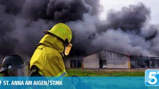 Ein Feuerwehrmann in gelber Uniform steht vor einem brennenden Gebäude, aus dem dicker Rauch aufsteigt. Ein weiterer Feuerwehrmann steht dahinter, beide tragen Helme. Der Ort ist St. Anna am Aigen/Stmk.