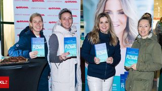 Vier Frauen stehen zusammen und halten Bücher mit dem Titel 'Die Magie des Flows'. Sie lächeln und scheinen für ein Foto zu posieren. Hinter ihnen ist ein großes Poster mit einer Frau mit blonden Haaren.