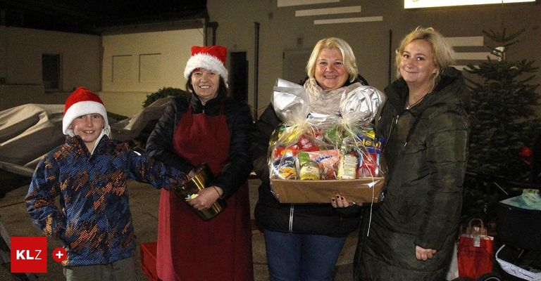 Drei Frauen und ein Kind stehen nachts zusammen und halten einen Korb mit Essen in der Hand und lächeln für die Kamera. Eine Frau trägt einen Weihnachtshut und eine andere hält einen Topf. Ein Gebäude und ein helles Licht sind im Hintergrund.