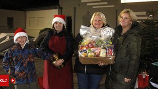 Drei Frauen und ein Kind stehen nachts zusammen und halten einen Korb mit Essen in der Hand und lächeln für die Kamera. Eine Frau trägt einen Weihnachtshut und eine andere hält einen Topf. Ein Gebäude und ein helles Licht sind im Hintergrund.