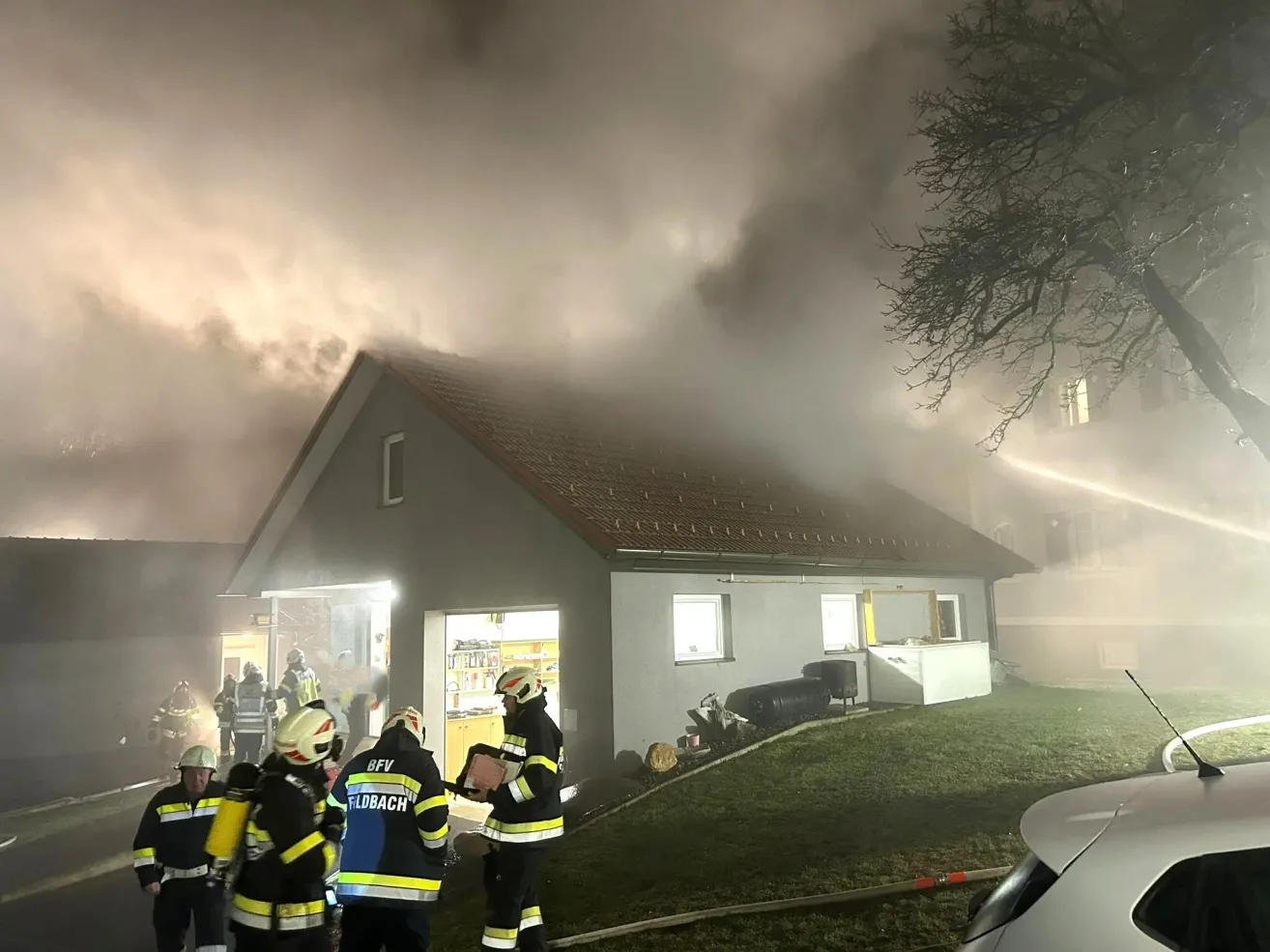 Feuerwehrleute bekämpfen ein Feuer in einem Haus. Rauch steigt vom Dach auf, und Feuerwehrleute verwenden Schläuche. Einige stehen vor dem Haus, andere in der Nähe eines Fahrzeugs.
