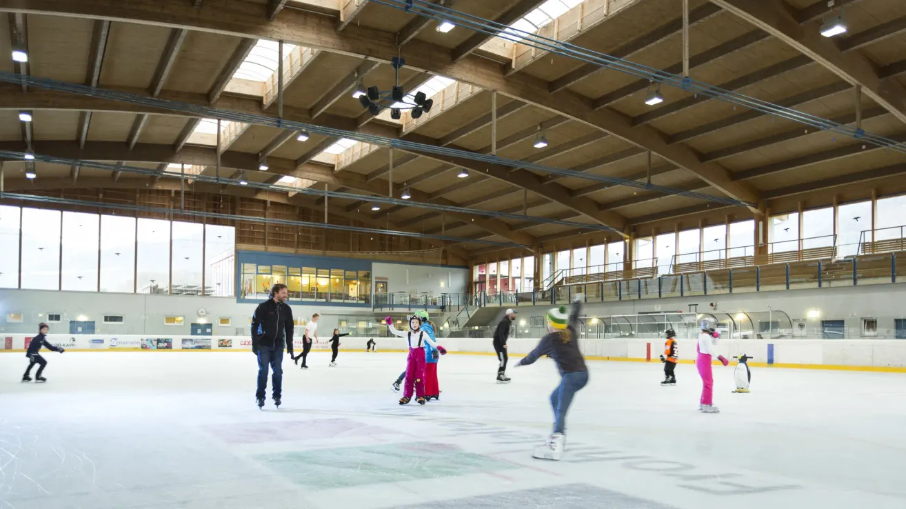 Eine überdachte Eislaufbahn mit mehreren Personen in Winterkleidung, die Schlittschuh laufen und einige stehen an der Seite. Die Bahn hat eine hohe Decke mit Holzbalken und Lampen.