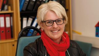 A woman with short blonde hair and glasses is smiling. She is wearing a red scarf and a black leather jacket. Behind her, a bookshelf with several binders is visible.