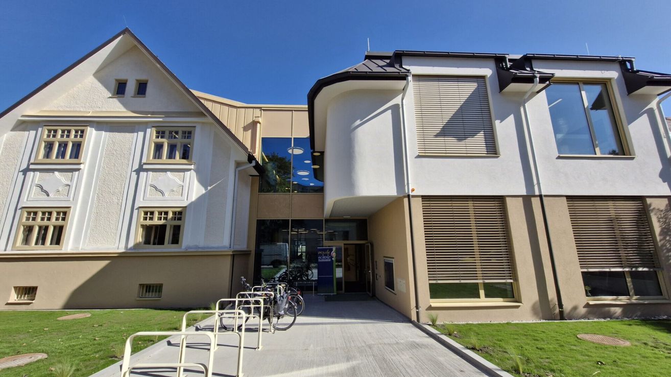 Der Eingang eines modernen Gebäudes mit geparkten Fahrrädern davor. Das Gebäude verfügt über Glaswände und einen blauen Banner.