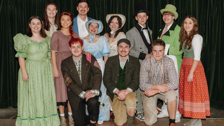 Eine Gruppe von Menschen in historischen Kostümen posiert für ein Foto. Sie lächeln und scheinen sich zu amüsieren. Der Hintergrund deutet auf eine Bühnenszene hin.