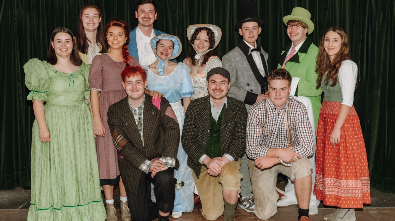 Eine Gruppe von Menschen in historischen Kostümen posiert für ein Foto. Sie lächeln und scheinen sich zu amüsieren. Der Hintergrund deutet auf eine Bühnenszene hin.