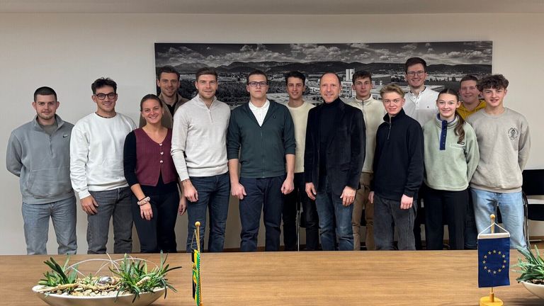 Eine Gruppe von Menschen posiert für ein Foto in einem Raum. Sie stehen vor einem Tisch. Eine Frau steht auf der linken Seite und ein Mann auf der rechten Seite. Ein Topf mit einer Pflanze steht auf dem Tisch. Hinter ihnen ist ein großes Foto an der Wand angebracht.