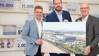 Drei Männer in Anzügen stehen vor einem großen Poster, das eine Fabrik zeigt. Die Fabrik ist mit Solarpaneelen ausgestattet und hat eine große Zahl 20 an der Wand.