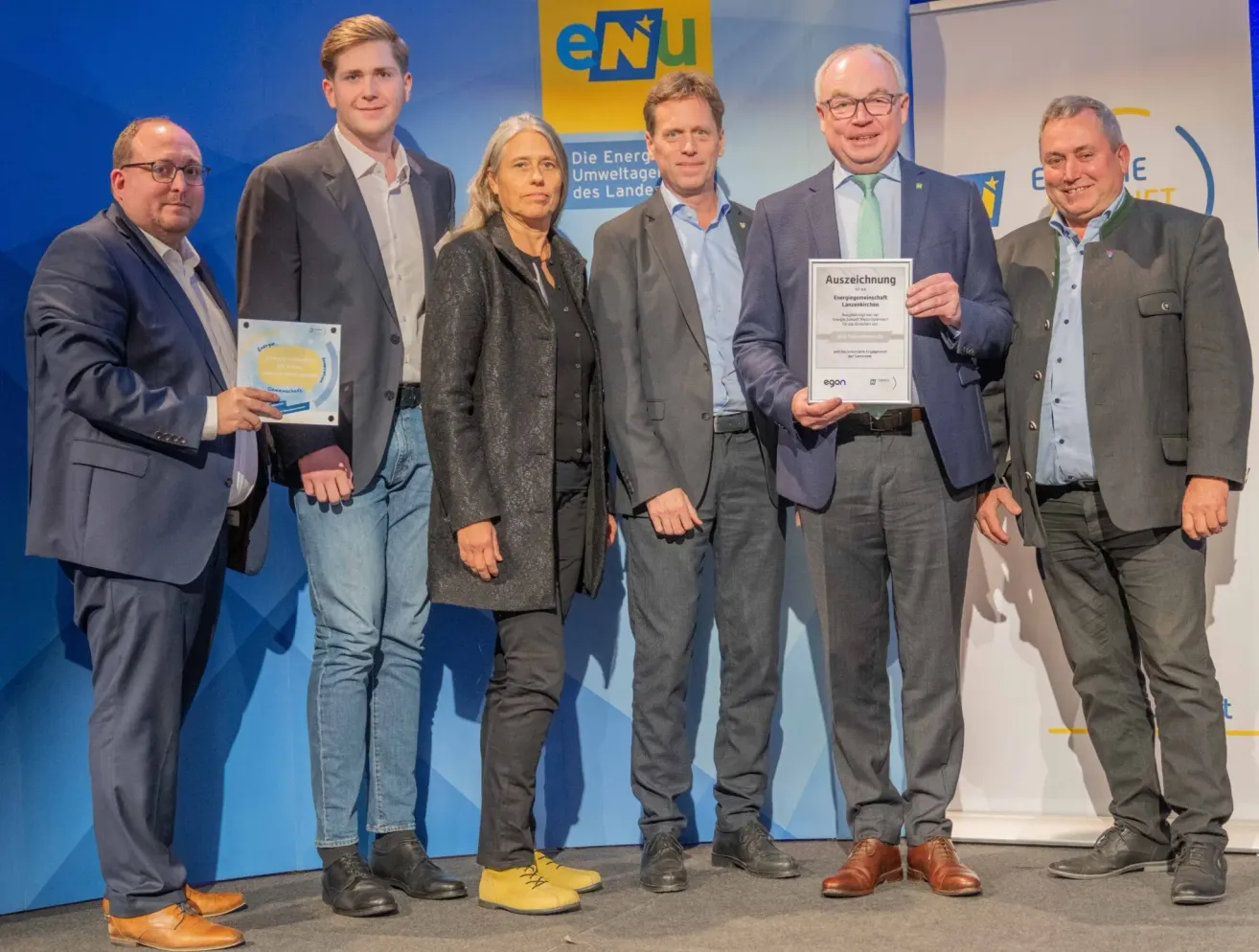 Sechs Personen in formeller Kleidung stehen auf einer Bühne, wahrscheinlich bei einer Preisverleihung. Ein Mann hält ein Zertifikat und steht neben einer Frau. Hinter ihnen zeigt ein Hintergrund ein gelbes und weißes Banner mit dem Text 'eNu'.
