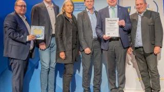 Sechs Personen in formeller Kleidung stehen auf einer Bühne, wahrscheinlich bei einer Preisverleihung. Ein Mann hält ein Zertifikat und steht neben einer Frau. Hinter ihnen zeigt ein Hintergrund ein gelbes und weißes Banner mit dem Text 'eNu'.