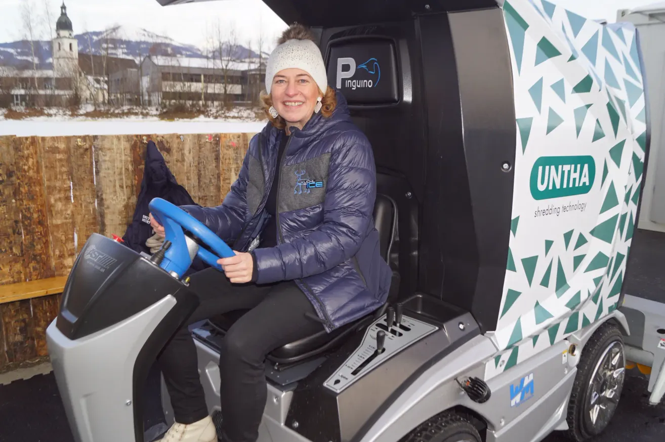 Eine Frau sitzt in einem Fahrzeug mit einem blauen Lenkrad und lächelt. Sie trägt eine weiße Mütze und eine blaue Jacke mit Logo. Hinter ihr ist ein Zaun und ein Gebäude in der verschneiten Landschaft zu sehen.