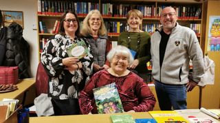Fünf Frauen und ein Mann stehen in einer Bibliothek. Sie alle lächeln und halten Bücher. Die Frau in der Mitte sitzt auf einem Stuhl und hält ein Buch mit deutschem Text.