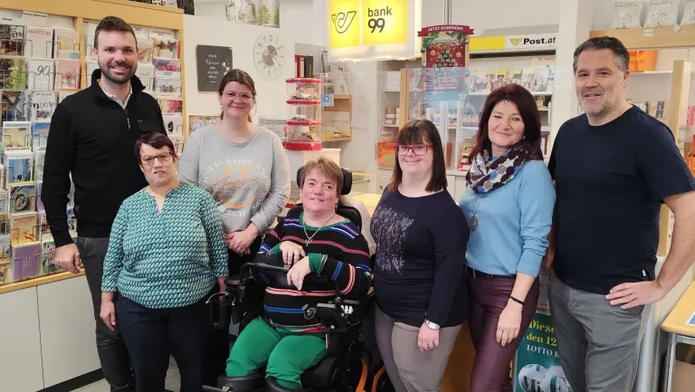 Five women are standing in a store, smiling and posing for a photo. One woman in a wheelchair is in the center. Behind them are shelves with products and a sign that reads 'bank 99'.