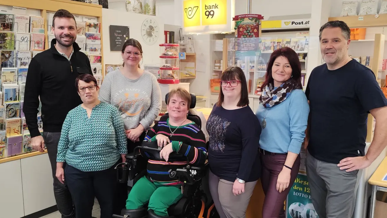 Fünf Frauen stehen in einem Geschäft, lächeln und posieren für ein Foto. Eine Frau im Rollstuhl steht in der Mitte. Hinter ihnen sind Regale mit Produkten und ein Schild, das 'Bank 99' steht.