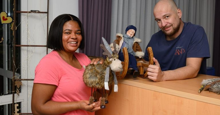 Eine Frau und ein Mann lächeln in die Kamera und halten beide Spielzeuge. Die Frau hält ein Spielzeugrentier und der Mann hält einen Spielzeugschlitten.