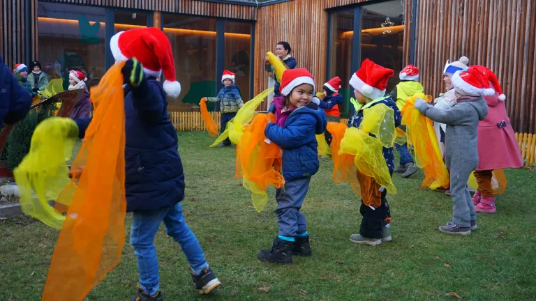 Eine Gruppe von Kindern, gekleidet in Winterkleidung und Weihnachtsmützen, tanzt auf einem Rasenfeld vor einem Gebäude im Hintergrund.