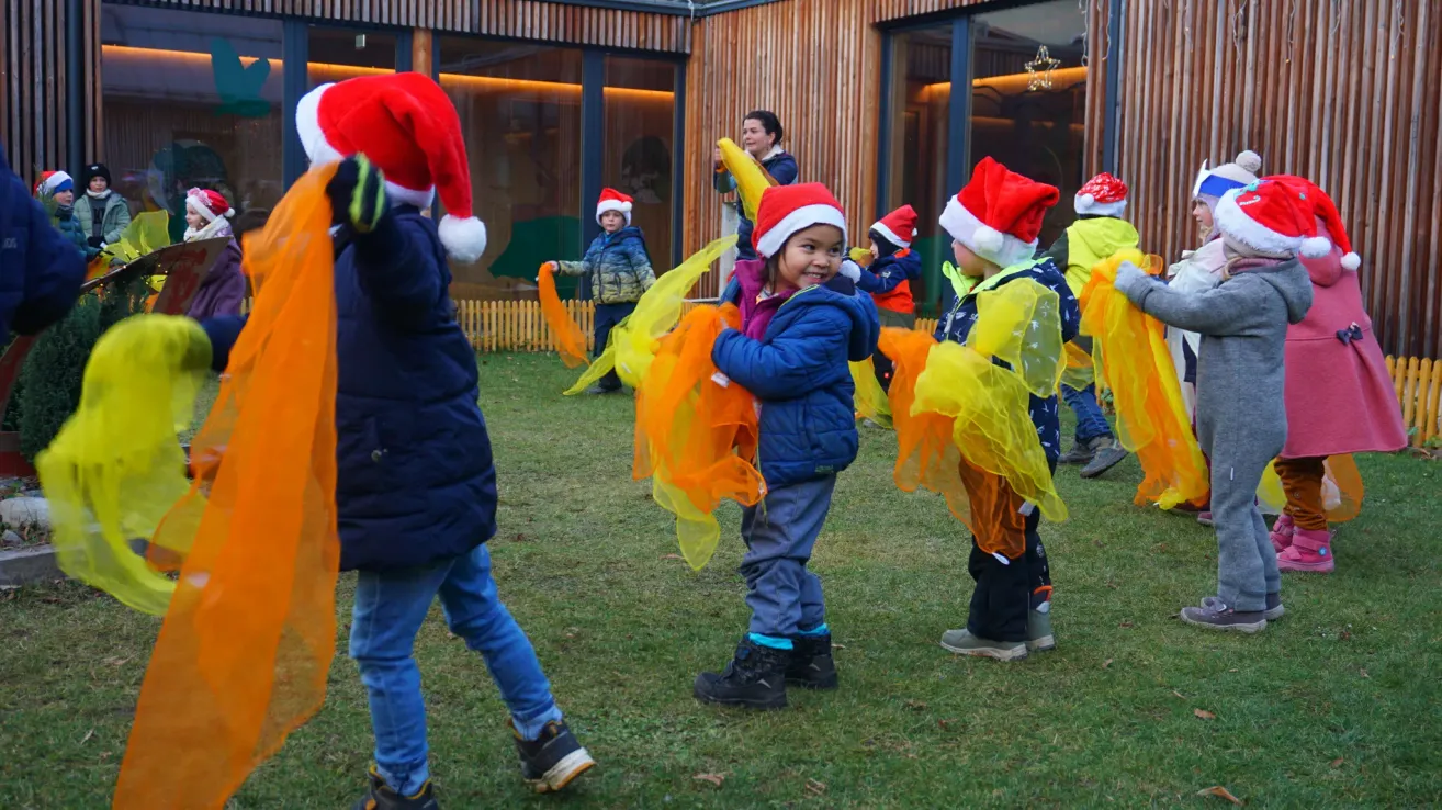 Eine Gruppe von Kindern, gekleidet in Winterkleidung und Weihnachtsmützen, tanzt auf einem Rasenfeld vor einem Gebäude im Hintergrund.