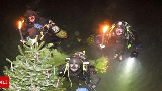 Three divers in scuba gear are underwater near a Christmas tree. They are illuminated by a light, and one holds a torch. The water is dark and green.