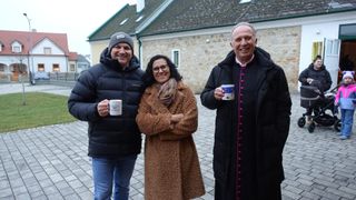 Drei Personen stehen vor einem Steingebäude und lächeln. Der Mann links trägt eine Mütze und hält eine Tasse. Die Frau in der Mitte hat eine Schürze und hält ihre Arme. Der Mann rechts hält eine Tasse und trägt einen Umhang.