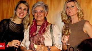 Three women are standing next to each other, smiling and holding wine glasses. The woman in the middle wears a red scarf, and the woman on the right wears a brown top. They are standing in front of a wooden wall.