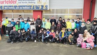 Eine Gruppe von Menschen in Winterkleidung posiert für ein Foto vor einem Gebäude mit einem Banner, das 'Willkommen zum Adventlauf mit Nordic Walking' besagt.