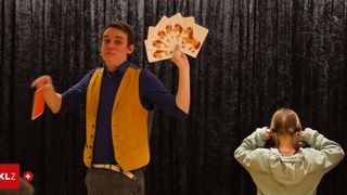 A man in a blue shirt and yellow vest is holding cards with horse images in front of a black curtain, while a young girl watches.