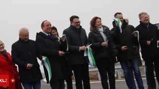 A group of people stands on a road, holding green and white ribbons, smiling for the camera, dressed in winter clothes.