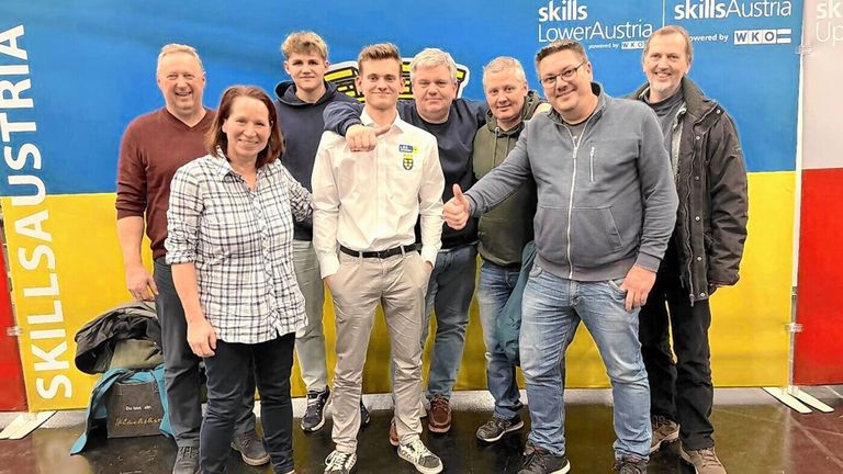 Eine Gruppe von Menschen steht zusammen vor einem Banner. Sie alle lächeln. Ein Mann zeigt mit dem Finger auf die Kamera.