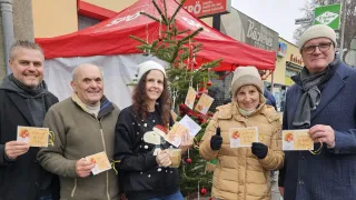 Three people stand in front of a decorated Christmas tree, smiling and holding cards. The man on the left holds a cup with a beverage. The woman in the center wears a Snoopy sweater. The woman on the right gives a thumbs up. Behind them is a red tent and a building with a sign.