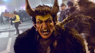 Eine Person in einem Hörnerkostüm läuft nachts auf der Straße. Hinter ihnen ist eine verschwommene Menge. Zwei Männer in Jacken stehen am Straßenrand.