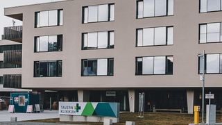 The exterior of a modern hospital building with glass windows, a sign reading 'Tauern Klinikum', and a grassy area in front.