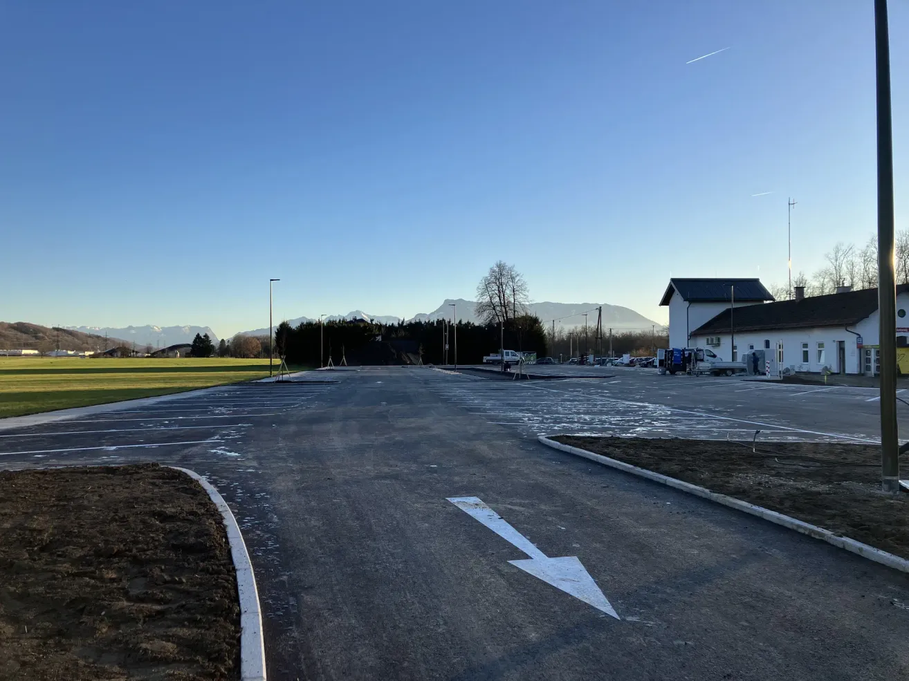 An empty parking lot with a white arrow on the road, a building, trees, and mountains in the background.