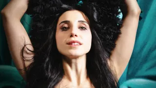 A woman with black hair wearing a black sequined dress poses against a teal background with black feathers on her head.