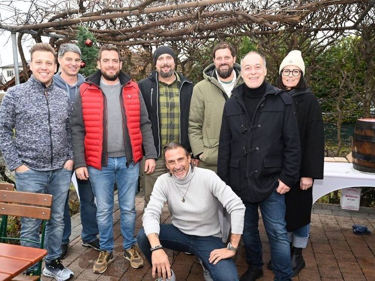 Eine Gruppe von Menschen, sowohl Männer als auch Frauen, stehen und sitzen draußen im Winter. Sie tragen warme Kleidung und scheinen für ein Foto zu posieren. Hinter ihnen steht ein Weihnachtsbaum und ein Tisch mit weißer Tischdecke.