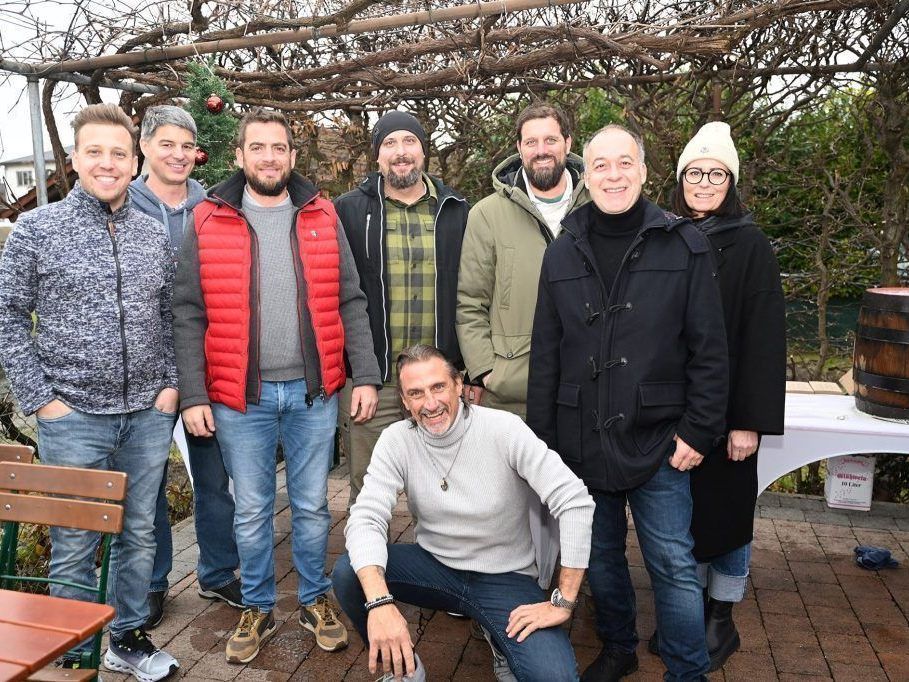 Eine Gruppe von Menschen, sowohl Männer als auch Frauen, stehen und sitzen draußen im Winter. Sie tragen warme Kleidung und scheinen für ein Foto zu posieren. Hinter ihnen steht ein Weihnachtsbaum und ein Tisch mit weißer Tischdecke.