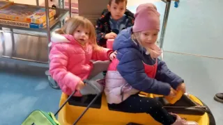 Three children sit in a yellow sled indoors, wearing winter coats. A green suitcase is nearby. Behind them, a table with toys and a shelf with books is visible.