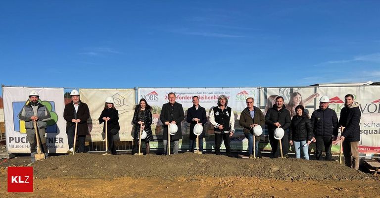 Eine Gruppe von Menschen steht in einer Reihe, Schaufeln in der Hand, an einer Baustelle. Hinter ihnen ist ein Banner mit Text und Bildern an einem Zaun befestigt.