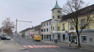 Eine Straße mit Gebäuden, einem Glockenturm, einem gelben Lieferwagen und Menschen, die auf dem Bürgersteig gehen.