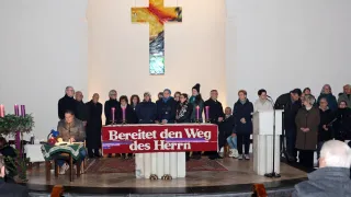 A group of people dressed in black stands in front of a church altar. The altar has a large cross on it. They are holding candles, and the sign says Bereitet den Weg des Herrn.