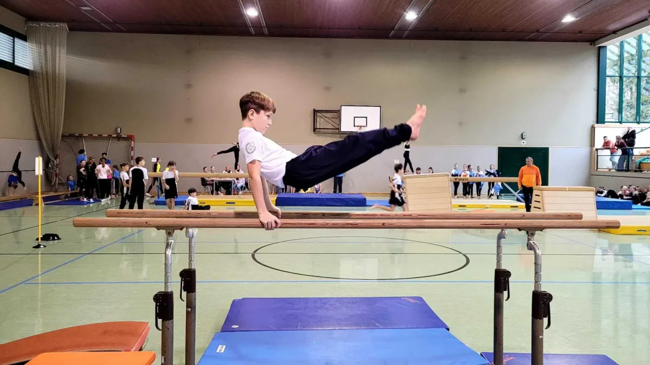 Ein junger Junge führt einen Handstand an einer Gymnastikstange in einer Turnhalle aus. Zuschauer sind anwesend, und es gibt verschiedene Geräte und einen Basketballkorb im Hintergrund.
